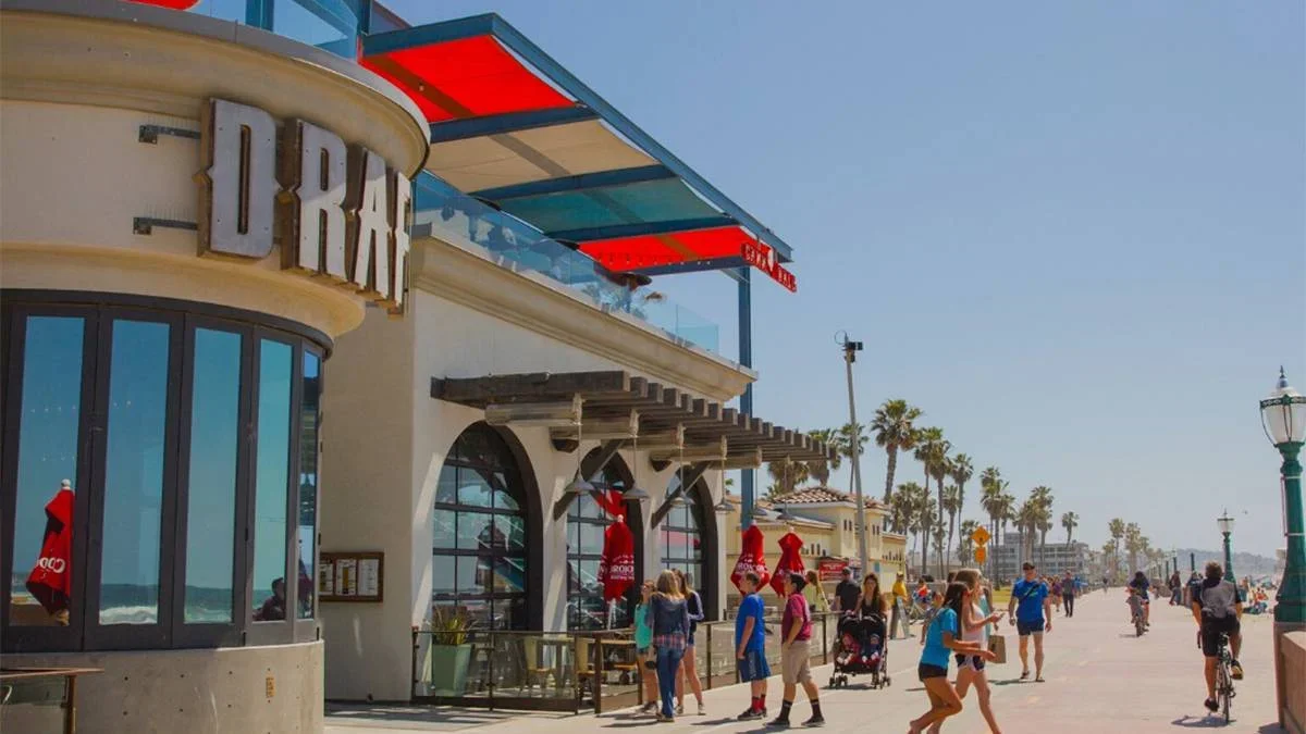 People scattered all over a sunny board walk with palm trees in the distance and building with a red white and blue awning at Belmont Park in San Diego, California